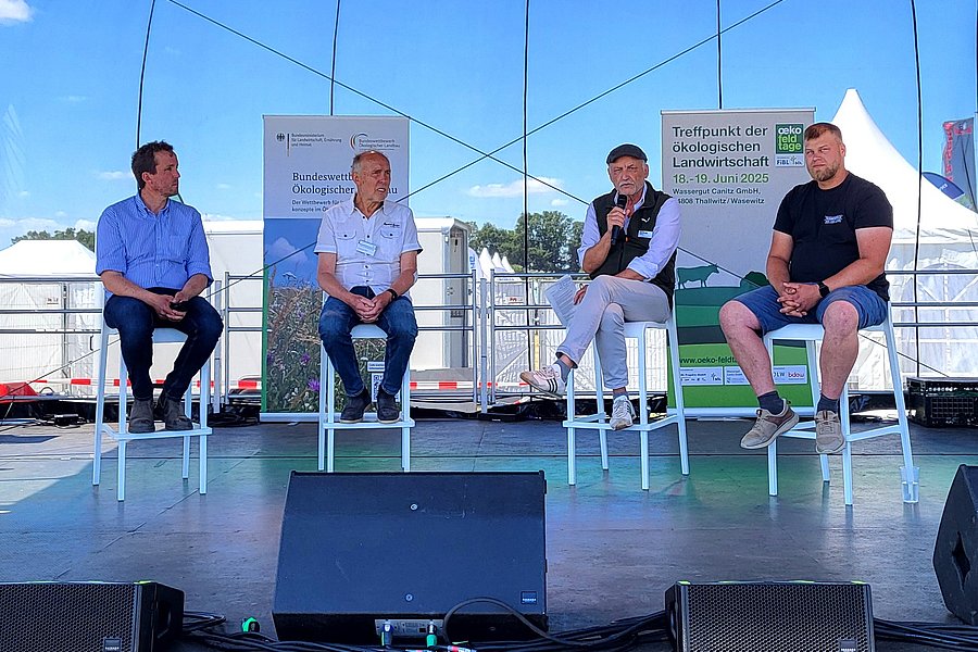 Vier Männer auf Stühlen sitzend auf einer Bühne während einer Podiumsdiskussion.