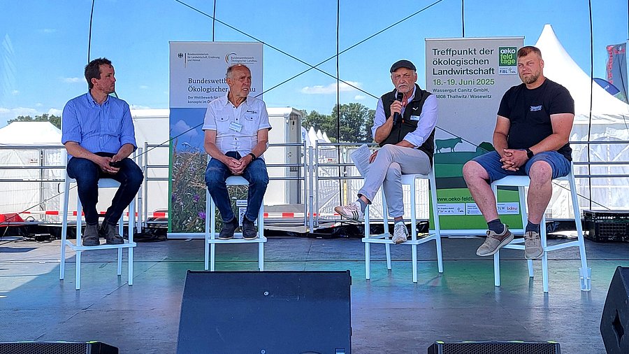 Vier Männer auf Stühlen sitzend auf einer Bühne während einer Podiumsdiskussion.